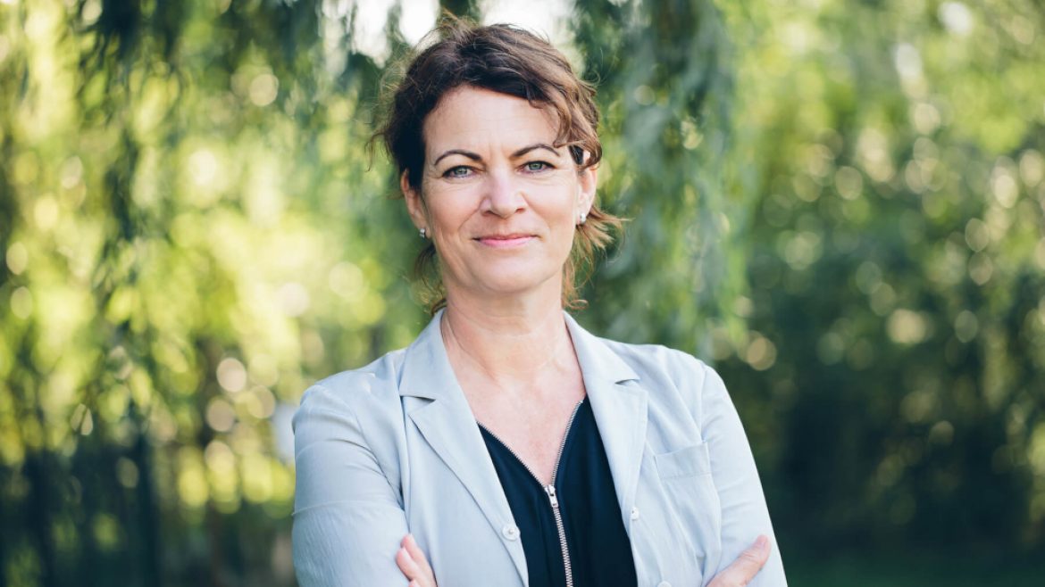 Eine Frau mit kurzen braunen Haaren in einem hellen Blazer steht im Freien mit verschränkten Armen und lächelt in die Kamera.