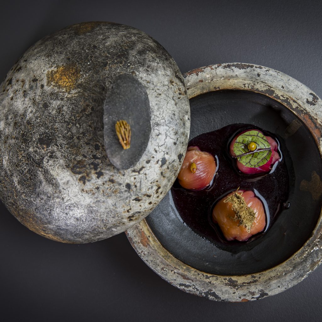 Eine rustikale Steinschale mit Deckel, in der sich drei kunstvoll angerichtete Knödel in einer dunklen Soße befinden.