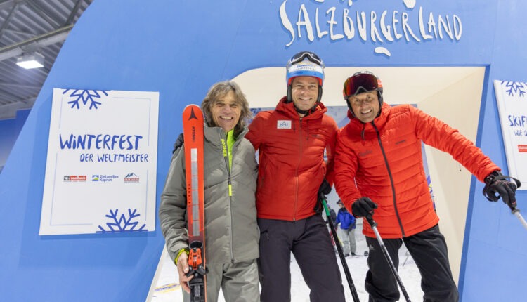 Drei lächelnde Männer in Winterkleidung posieren mit Skiern vor einem blauen SalzburgerLand-Bogen bei einer Skiveranstaltung.