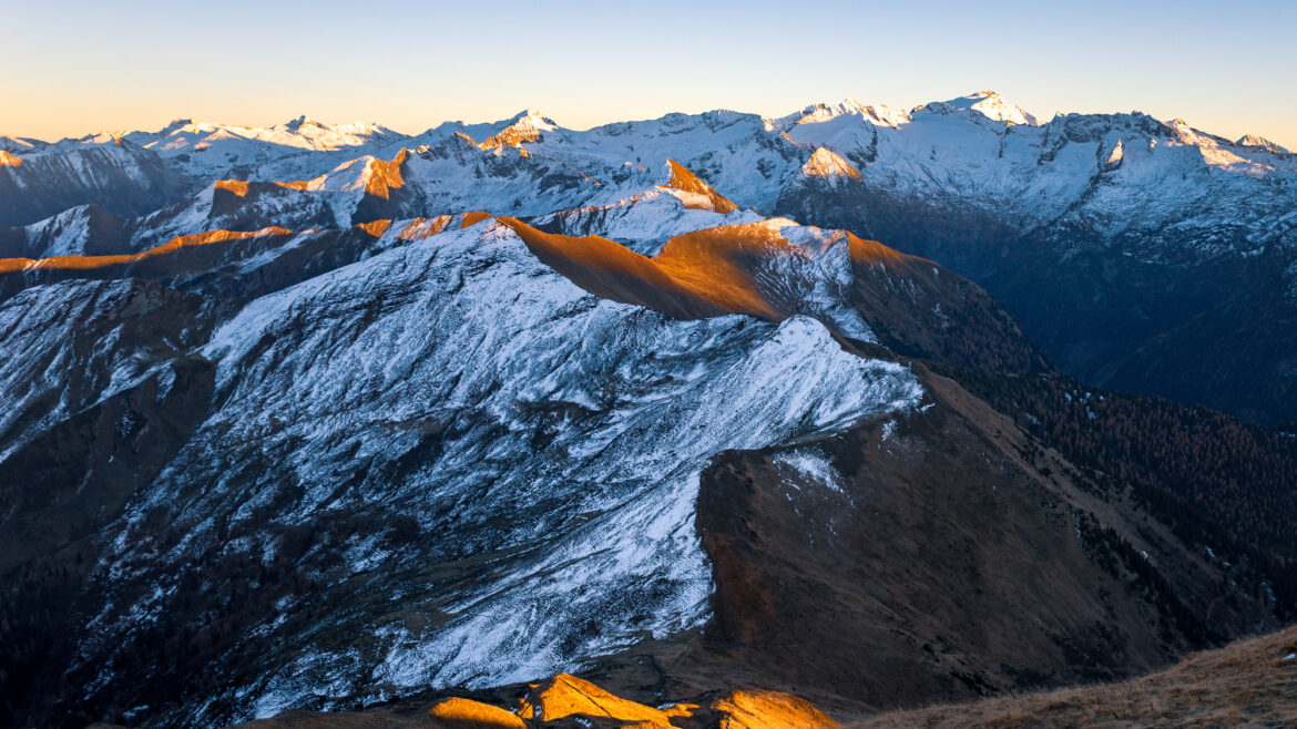 Verschneite Berggipfel bei Sonnenaufgang mit goldenem Licht, das die Bergkämme und fernen Berge im Hintergrund hervorhebt.