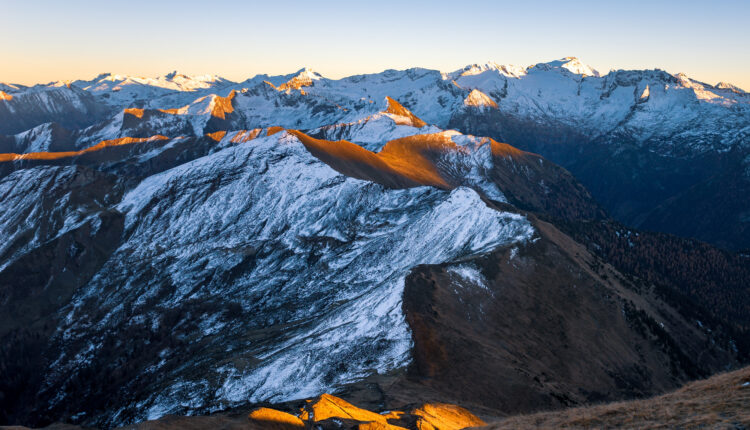 Verschneite Berggipfel bei Sonnenaufgang mit goldenem Licht, das die Bergkämme und fernen Berge im Hintergrund hervorhebt.