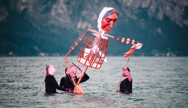 Drei Personen im Wasser bedienen eine große Puppe mit bemaltem Gesicht und Holzrahmen, im Hintergrund Berge.