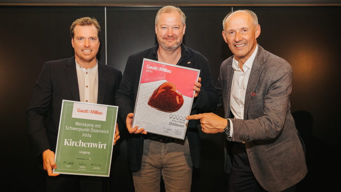 Drei Männer stehen lächelnd da, halten und zeigen auf die Gault&Millau 2024 Restaurant- und Kuchenpreise.