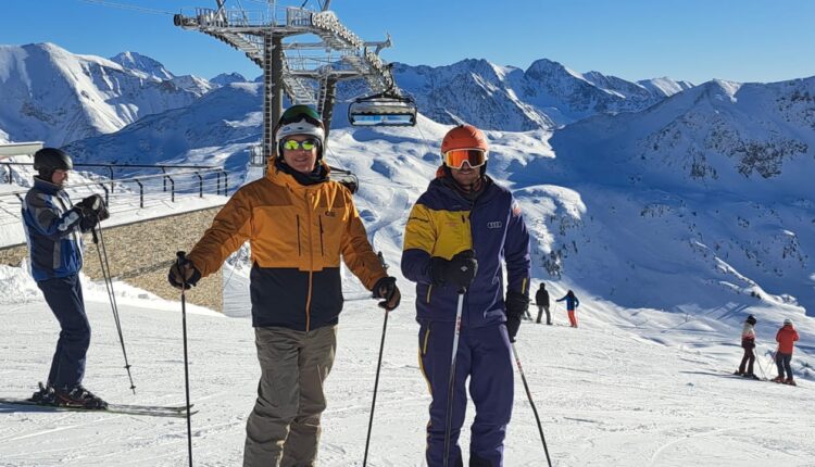 Zwei Skifahrer posieren an einem sonnigen Tag auf einer verschneiten Piste mit Bergen und einem Skilift im Hintergrund.