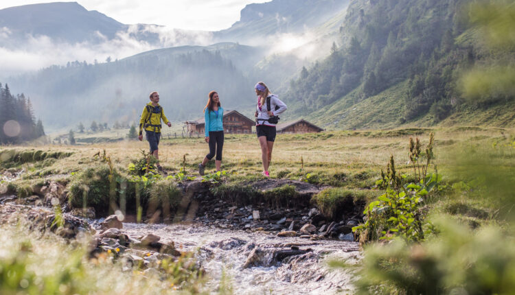 Drei Personen wandern entlang eines Baches in einer grasbewachsenen Berglandschaft mit Nebel und Hütten im Hintergrund.