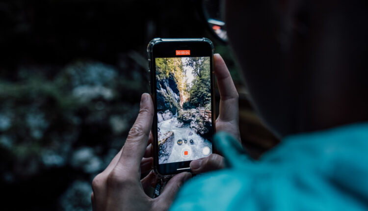 Eine Person fotografiert eine Wald- und Flussszene mit einem Smartphone in beiden Händen.