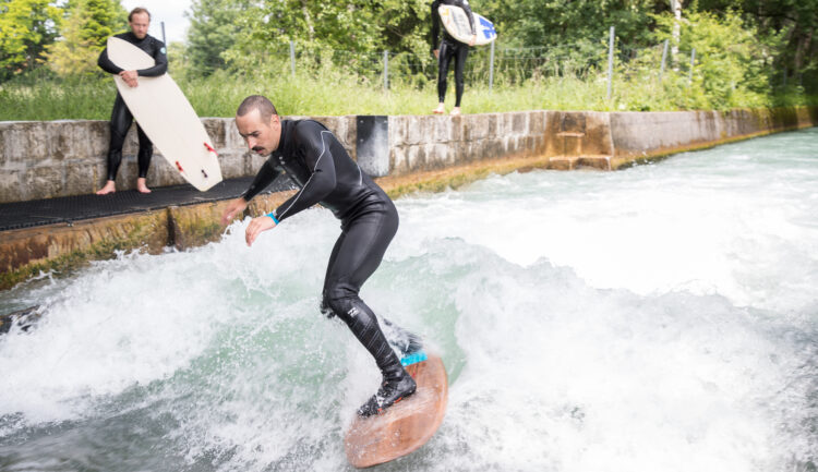 Ein Mann surft auf einer Flusswelle, während zwei Personen mit Surfbrettern vom Betonufer aus zusehen.