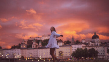 Frau in einem weißen Kleid dreht sich fröhlich im Freien bei Sonnenuntergang mit einer Stadt und einem dramatischen Himmel im Hintergrund.