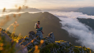 Ein Wanderer sitzt auf einem Felsvorsprung und blickt auf neblige Berge und ein Tal voller Wolken bei Sonnenaufgang.