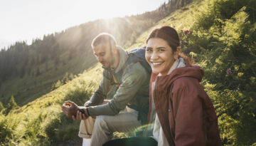 Zwei Menschen sitzen auf einem grasbewachsenen Hügel im Sonnenlicht; der eine lächelt in die Kamera, der andere überprüft seine Smartwatch.