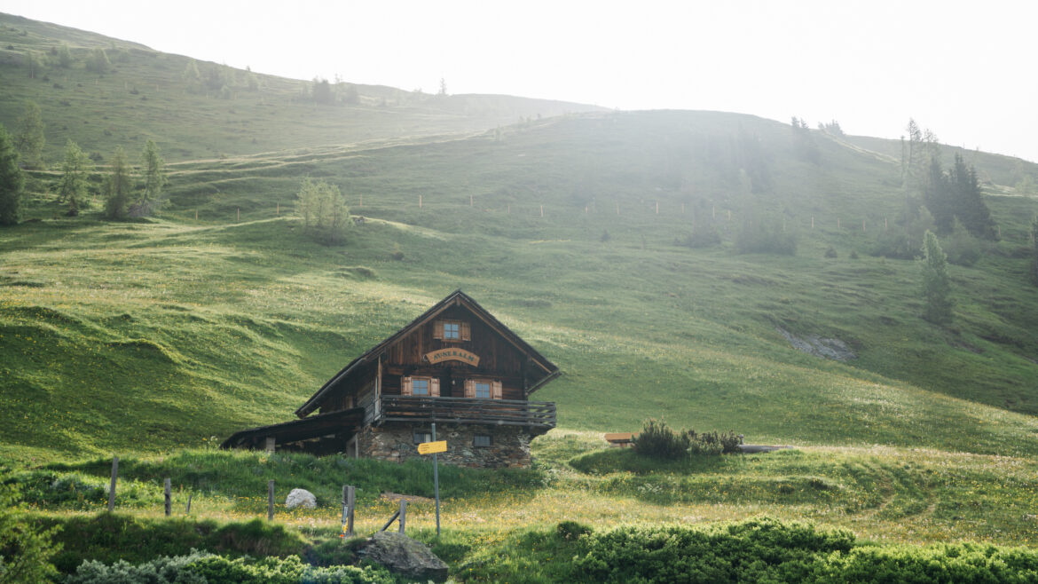 Eine Holzhütte steht auf einem grasbewachsenen Hügel, umgeben von grünen Feldern und Bäumen unter einem strahlenden Himmel.