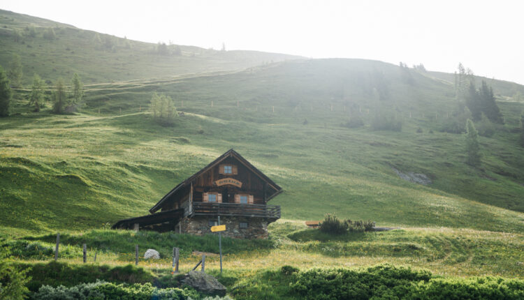 Eine Holzhütte steht auf einem grasbewachsenen Hügel, umgeben von grünen Feldern und Bäumen unter einem strahlenden Himmel.