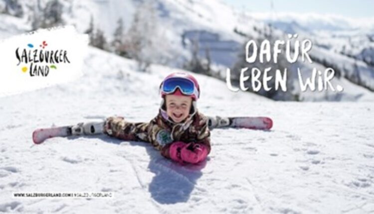 Ein lächelndes Kind in Skiausrüstung macht einen Spagat auf einer verschneiten Piste, mit Bergen im Hintergrund.