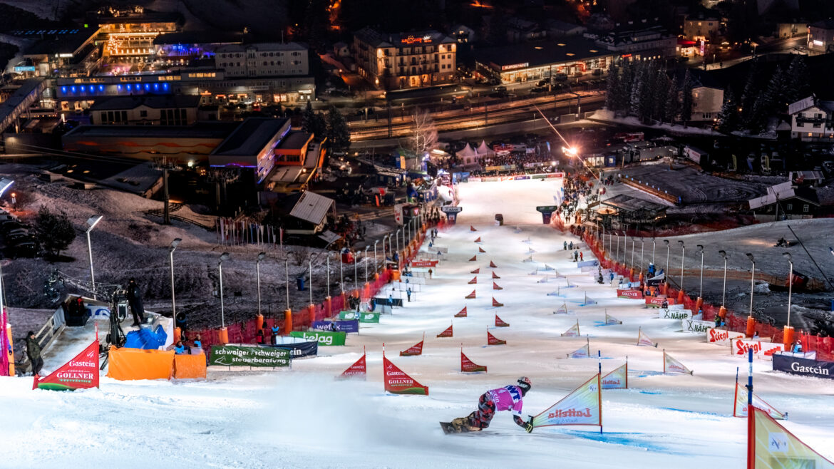 Ein Snowboarder fährt nachts eine Slalomstrecke hinunter, im Hintergrund sind Lichter und Gebäude zu sehen.