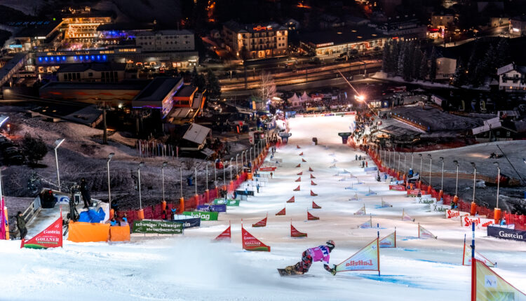 Ein Snowboarder fährt nachts eine Slalomstrecke hinunter, im Hintergrund sind Lichter und Gebäude zu sehen.