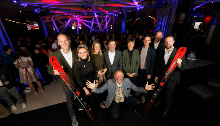Eine Gruppe von Menschen posiert und lächelt bei einer Indoor-Veranstaltung, zwei halten rote Skier, im Hintergrund eine Menschenmenge.