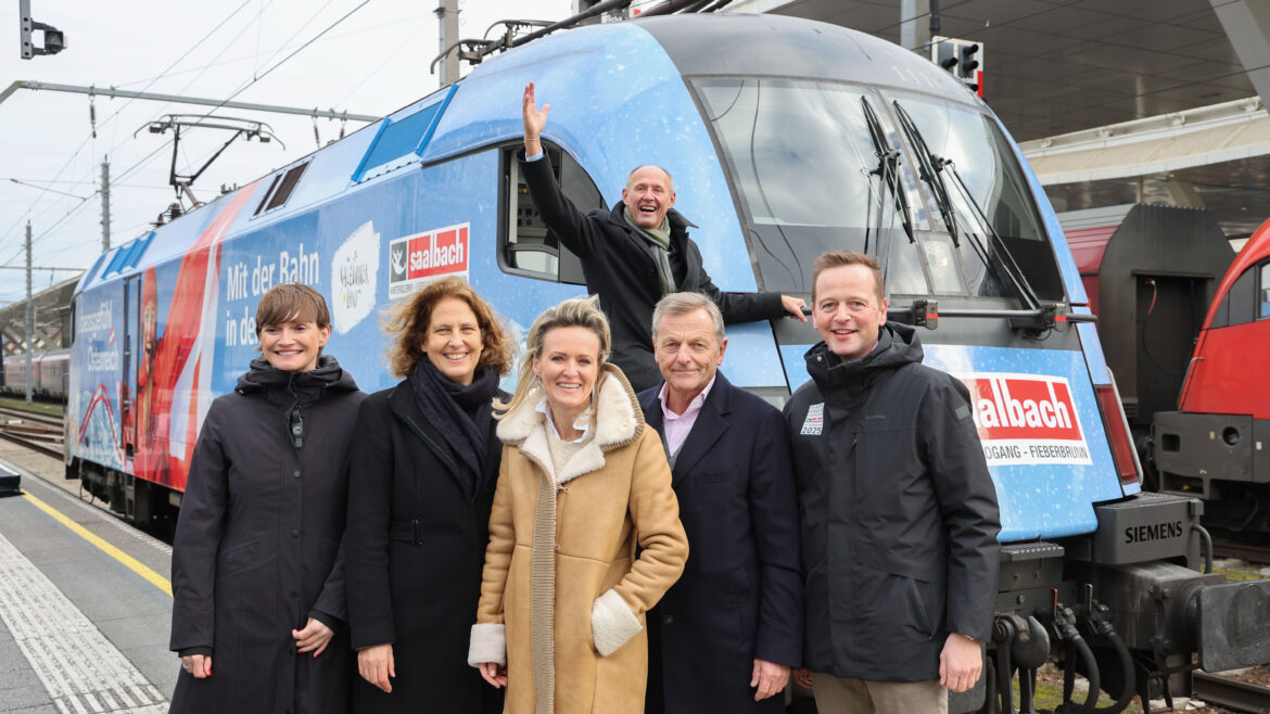 Sechs Personen posieren und lächeln vor einem blauen Zug; eine Person winkt aus dem Führerstand des Zuges.