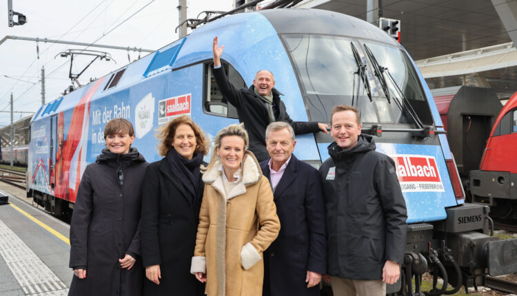 Sechs Personen posieren und lächeln vor einem blauen Zug; eine Person winkt aus dem Führerstand des Zuges.