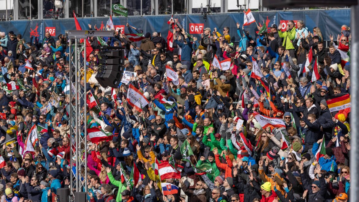 Eine große Menschenmenge auf den Tribünen schwenkt Fahnen und jubelt bei einer Veranstaltung im Freien, viele tragen bunte Jacken und Hüte.