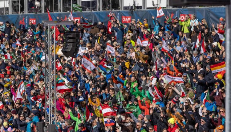 Eine große Menschenmenge auf den Tribünen schwenkt Fahnen und jubelt bei einer Veranstaltung im Freien, viele tragen bunte Jacken und Hüte.