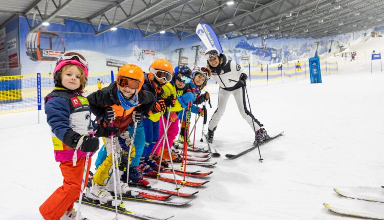 Ein Skilehrer posiert mit einer Gruppe von Kindern in farbenfroher Ausrüstung auf einer Indoor-Skipiste.