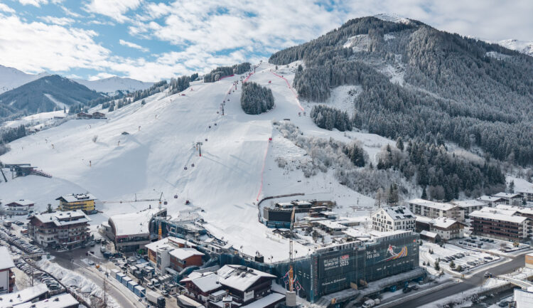 Ein verschneites Skigebiet mit Sesselliften, Pisten und Gebäuden am Fuße eines bewaldeten Berges.