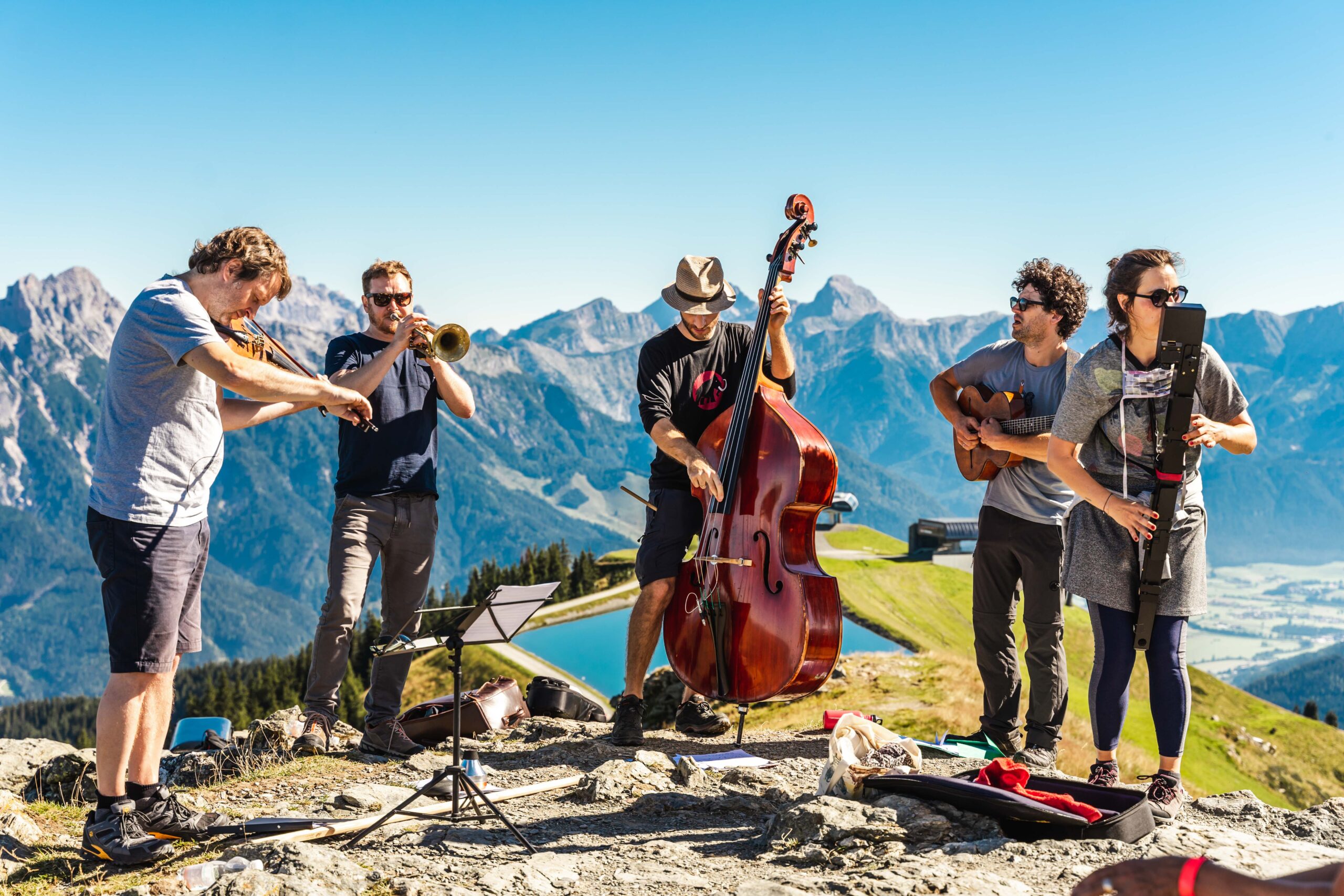 Fünf Musiker spielen Instrumente im Freien auf einem Berg mit blauem Himmel und malerischen Gipfeln im Hintergrund.