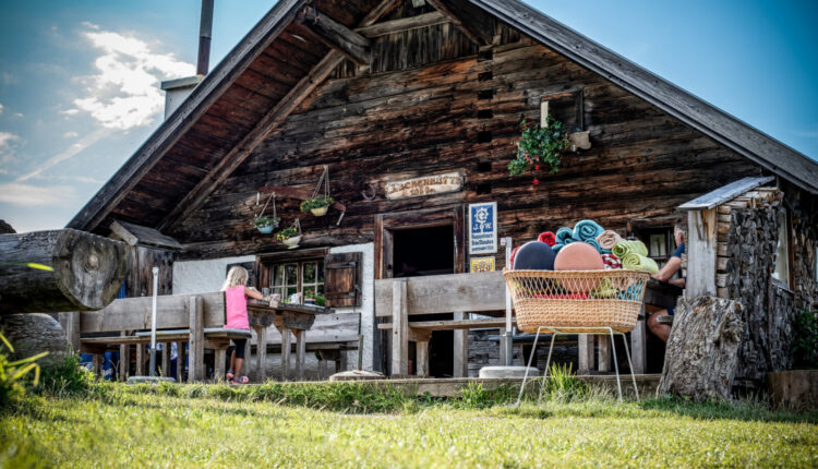 Eine rustikale Holzhütte mit Sitzgelegenheiten im Freien und Menschen, die sich an einem sonnigen Tag entspannen, gesehen von einer Wiese aus.