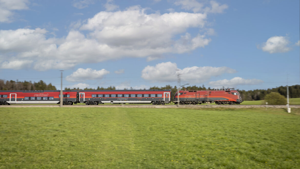 Ein roter Personenzug fährt schnell durch ein grünes Feld unter einem teilweise bewölkten Himmel.