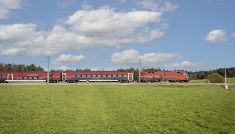 Ein roter Personenzug fährt schnell durch ein grünes Feld unter einem teilweise bewölkten Himmel.