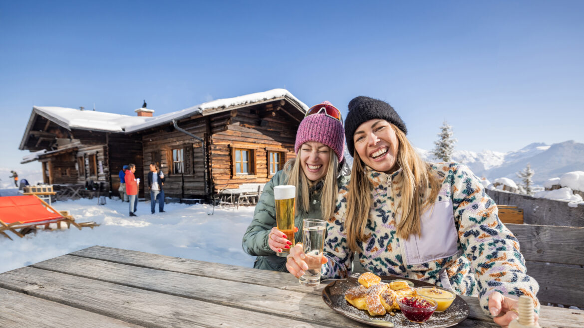 Zwei Frauen in Winterkleidung lachen und essen an einem Holztisch im Freien bei einer verschneiten Hütte in den Bergen.