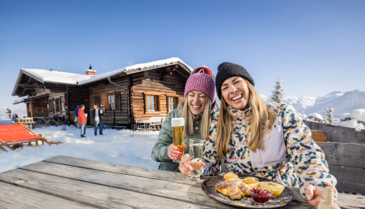 Zwei Frauen in Winterkleidung lachen und essen an einem Holztisch im Freien bei einer verschneiten Hütte in den Bergen.