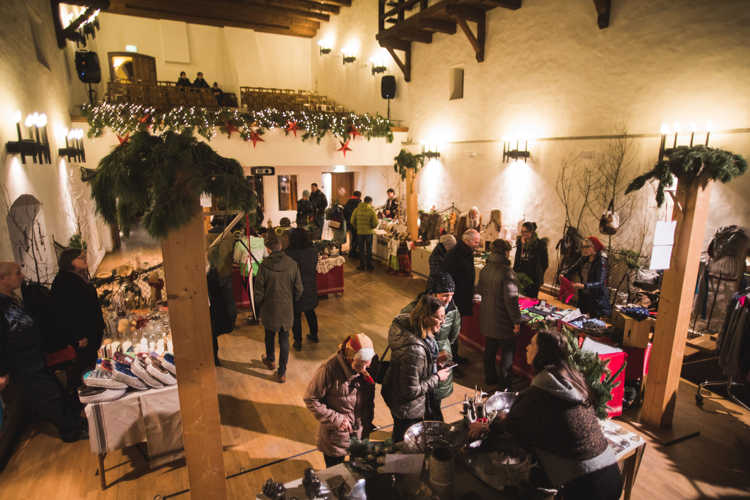 Weihnachtsmarkt in einer Halle mit festlichen Ständen, Dekorationen und Lichterketten auf einem Balkon.