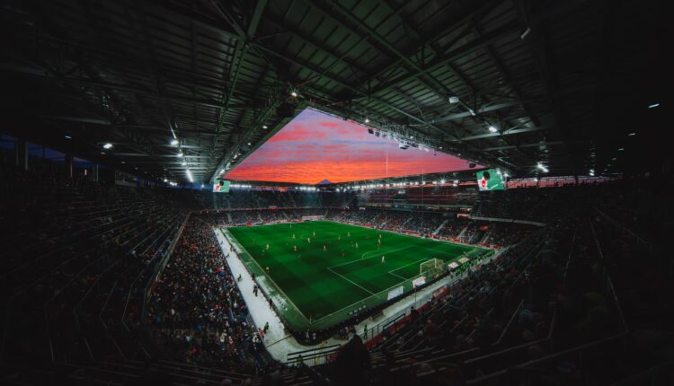 Ein vollbesetztes Stadion ist Schauplatz eines Fußballspiels unter hellen Lichtern und einem farbenprächtigen Sonnenuntergang am Himmel.