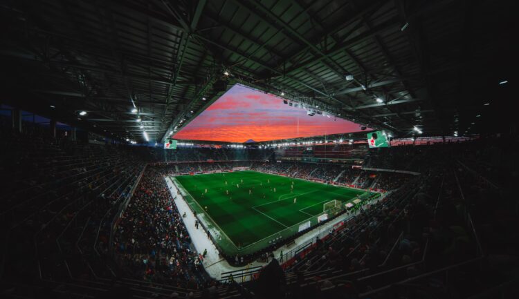Ein vollbesetztes Stadion ist Schauplatz eines Fußballspiels unter hellen Lichtern und einem farbenprächtigen Sonnenuntergang am Himmel.