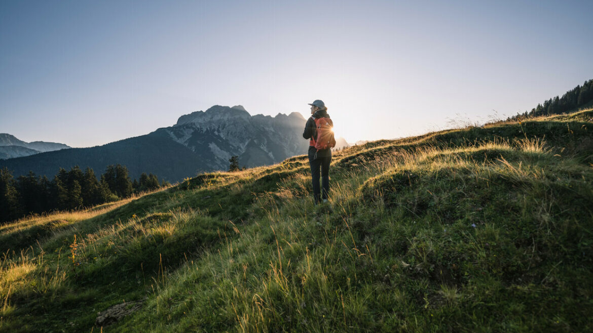 Eine Person wandert bei Sonnenaufgang auf einem grasbewachsenen Hügel, mit Bergen und Bäumen im Hintergrund.