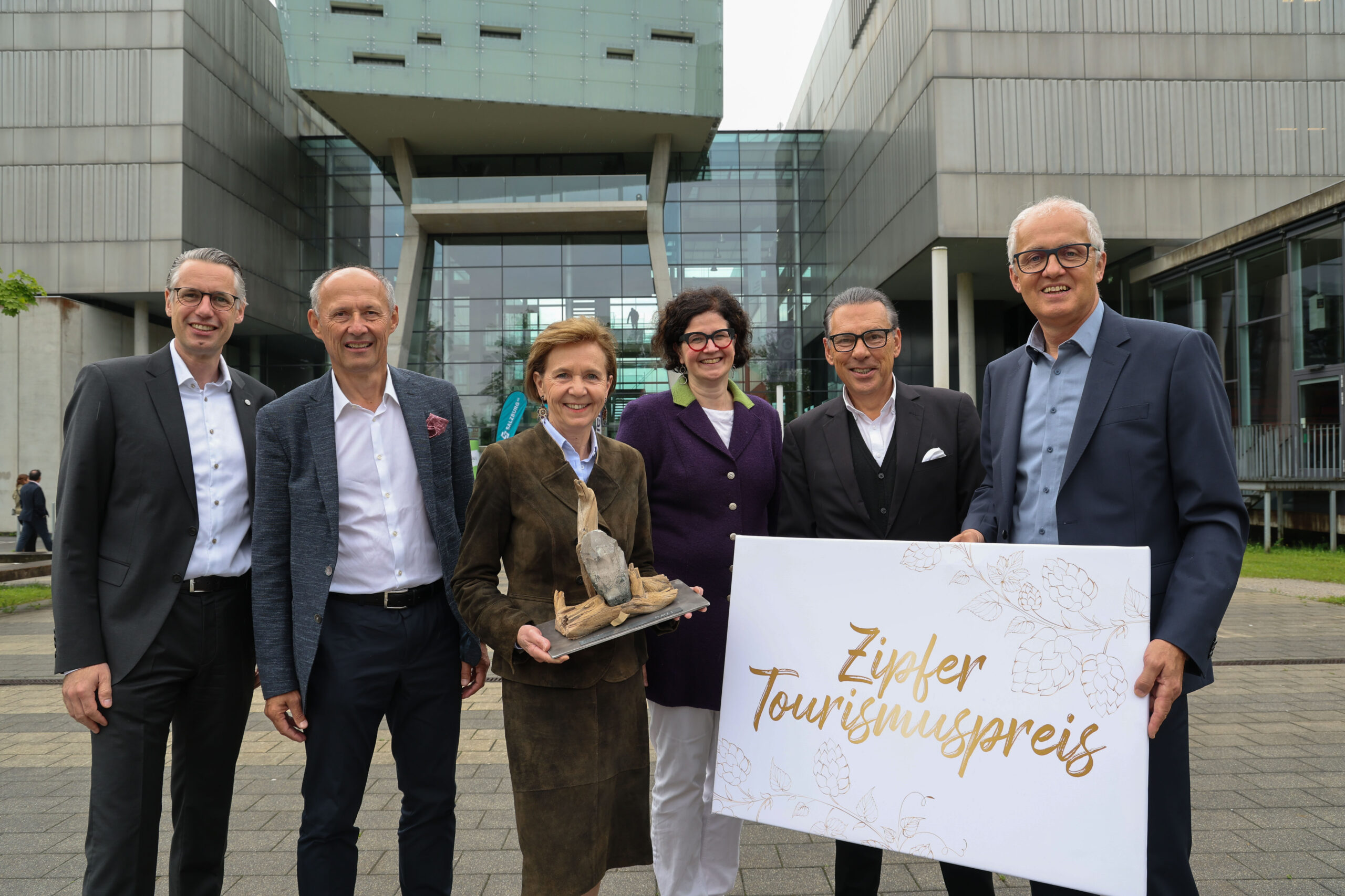 Sechs Personen stehen vor einem modernen Gebäude und halten eine Trophäe und ein Schild mit der Aufschrift Zipfer Tourismuspreis in der Hand.