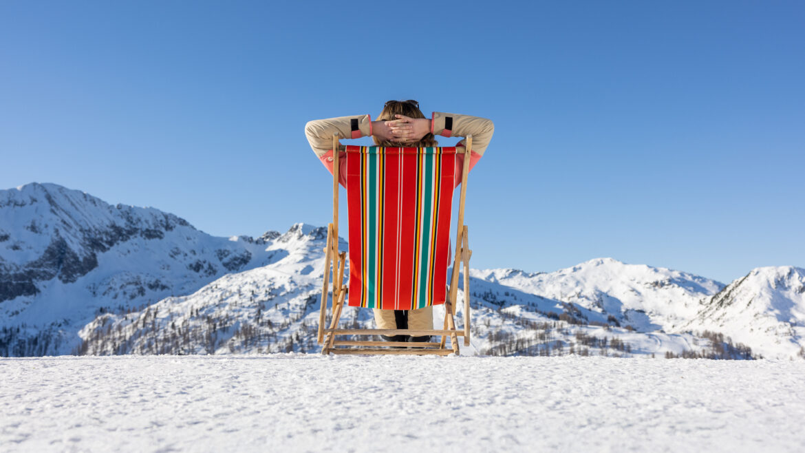 Person, die sich in einem gestreiften Liegestuhl auf verschneitem Boden entspannt, mit Blick auf schneebedeckte Berge unter einem klaren blauen Himmel.