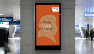 Digitales Plakat in einem Bahnhof mit Mozarts Silhouette und Text: Salzburg feiert das Genie auf orangem Hintergrund.