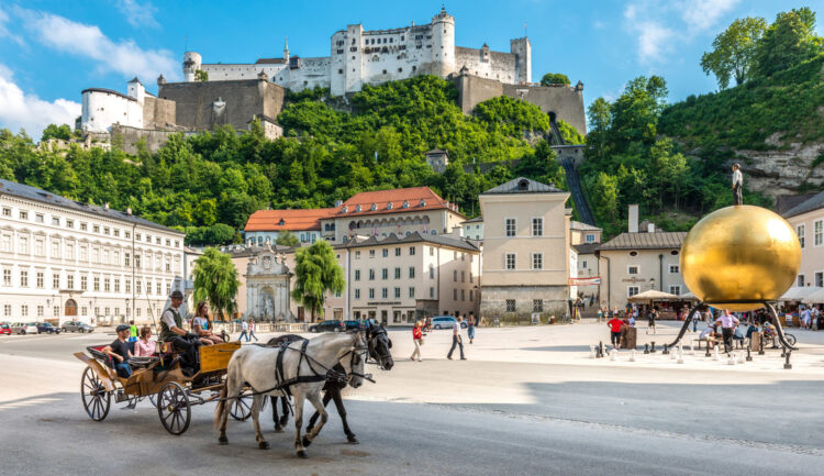 Eine Pferdekutsche auf einem sonnigen Stadtplatz mit einer Festung auf einem Hügel und einer großen goldenen Kugelskulptur.