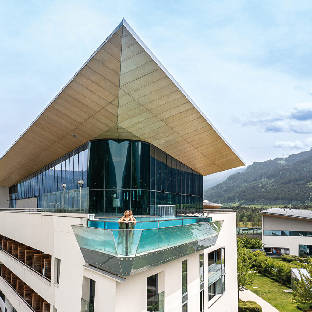 Modernes gläsernes Gebäude mit Spitzdach vor einer Bergkulisse; Person steht auf einem Balkon.
