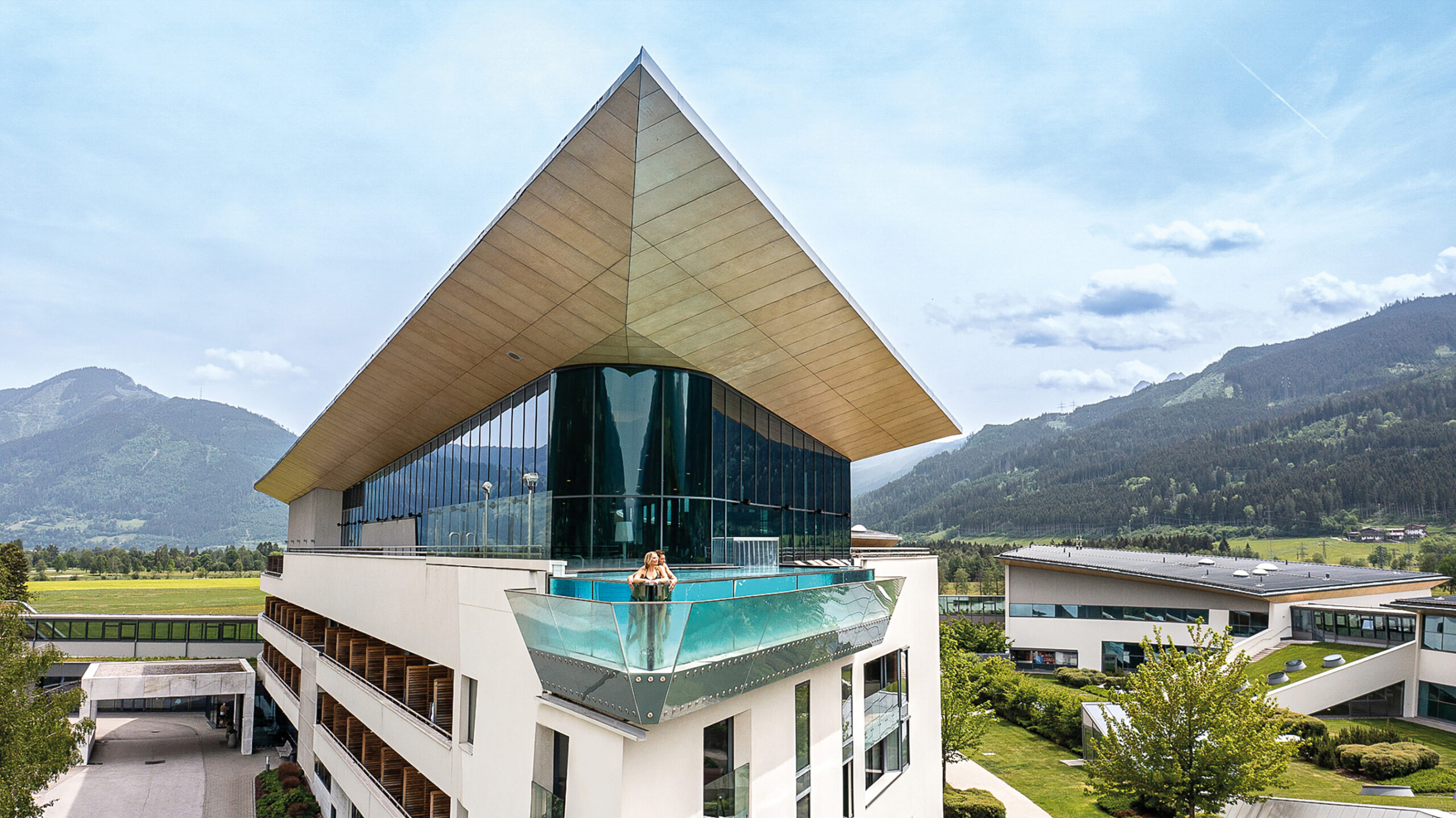 Modernes gläsernes Gebäude mit Spitzdach vor einer Bergkulisse; Person steht auf einem Balkon.