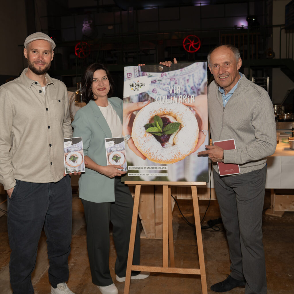 Drei Personen stehen in einem Raum und halten ein großes Lebensmittelplakat und Broschüren in der Hand, während im Hintergrund ein Esszimmer eingerichtet ist.