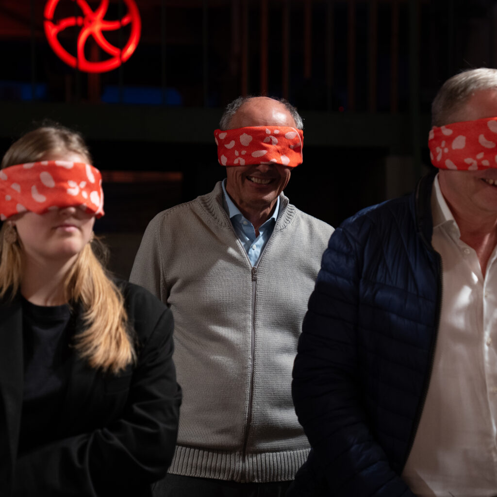 Drei Personen, die rote Augenbinden mit weißen Punkten tragen, stehen in einem Raum und lächeln.