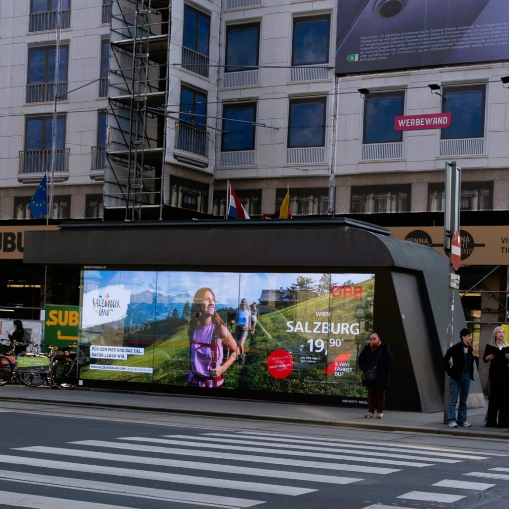 Eine belebte Straße in der Stadt mit einer großen digitalen Werbetafel, die eine Frau beim Wandern in Salzburg und verschiedene Personen in der Nähe zeigt.