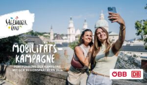Zwei Frauen lächeln und machen ein Selfie im Freien mit einer Stadtlandschaft und Kuppelgebäuden im Hintergrund.