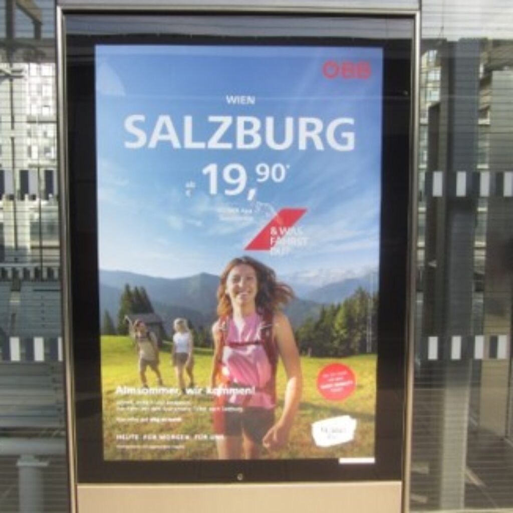 Zugwerbung nach Salzburg (19,90 Euro) mit lächelnden Wanderern in einer sonnigen Berglandschaft, auf einem Bahnsteig.
