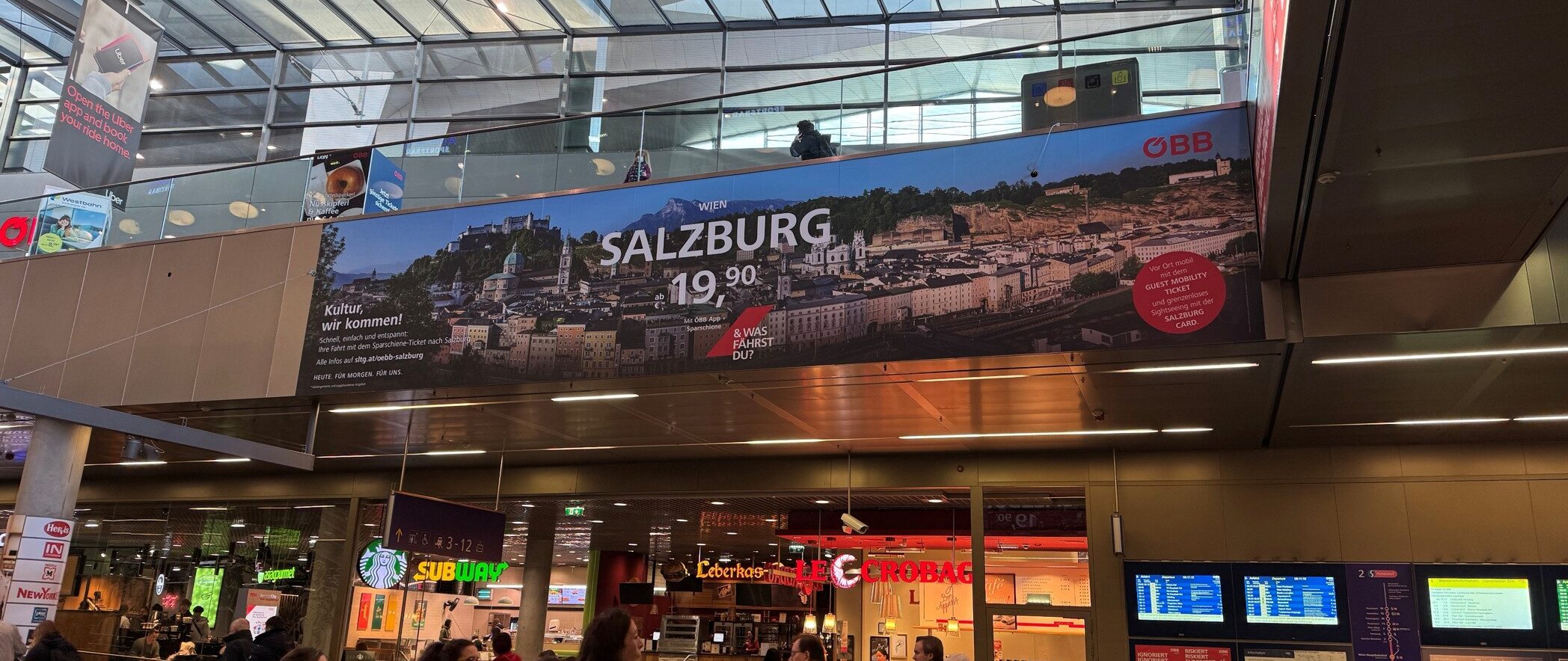 Große Salzburger Reisewerbung über dem belebten Bahnhofsinneren mit darunter sichtbaren Menschen und Geschäften.