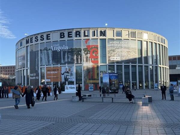Rund um das Gebäude der Messe Berlin mit großen Bannern, spazieren gehende und sich versammelnde Menschen unter einem klaren blauen Himmel.
