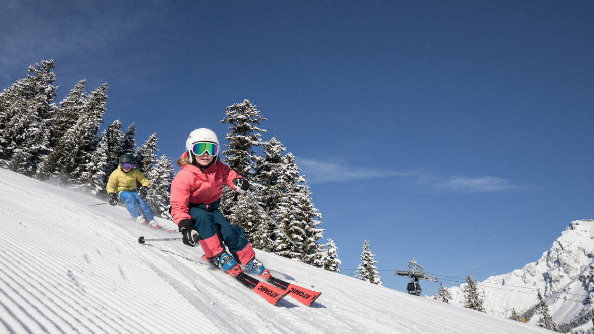 Zwei Personen fahren auf einer präparierten, verschneiten Piste mit Bäumen und Skilift im Hintergrund unter blauem Himmel Ski.